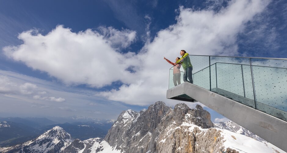 Two people stand on a glass viewing platform above snow-covered cliffs of the Dachstein massif | © Harald Steiner