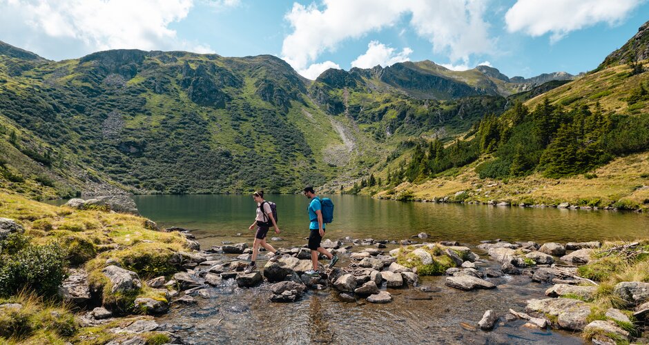 Two hikers with backpacks cross a stream by a mountain lake with green mountains and a blue sky behind | © Mathäus Gartner