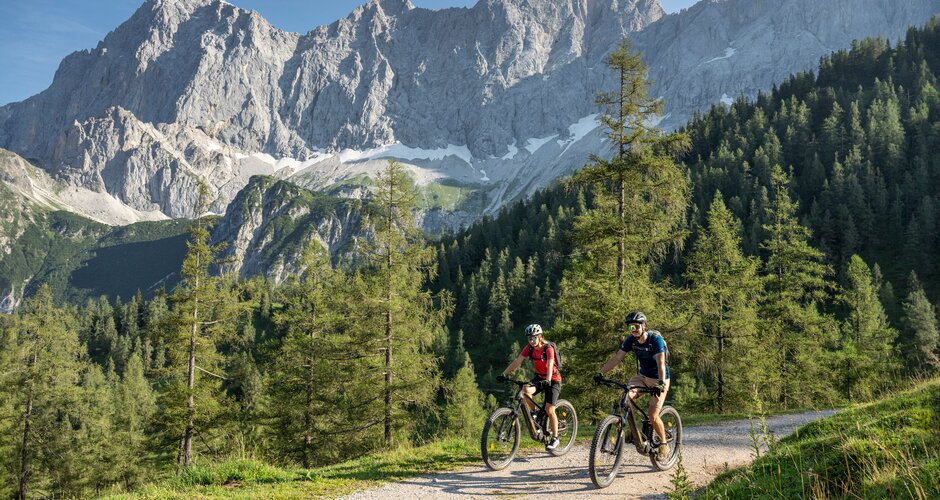 Two mountain bikers ride along a gravel path through alpine terrain with forest and a bright rock face below a blue sky | © Lorenz Masser