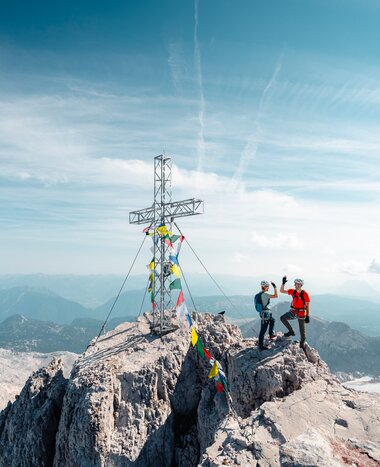 Two climbers wearing helmets stand beside a summit cross on a rocky peak above a wide alpine landscape | © Mathäus Gartner