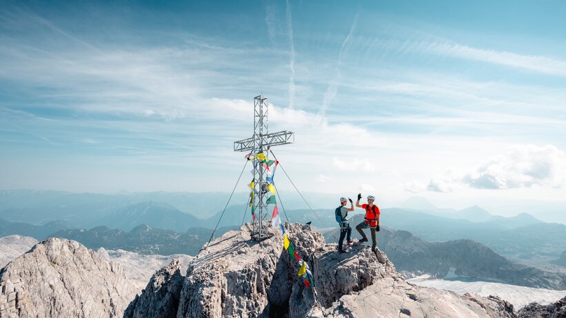 Two climbers wearing helmets stand beside a summit cross on a rocky peak above a wide alpine landscape | © Mathäus Gartner