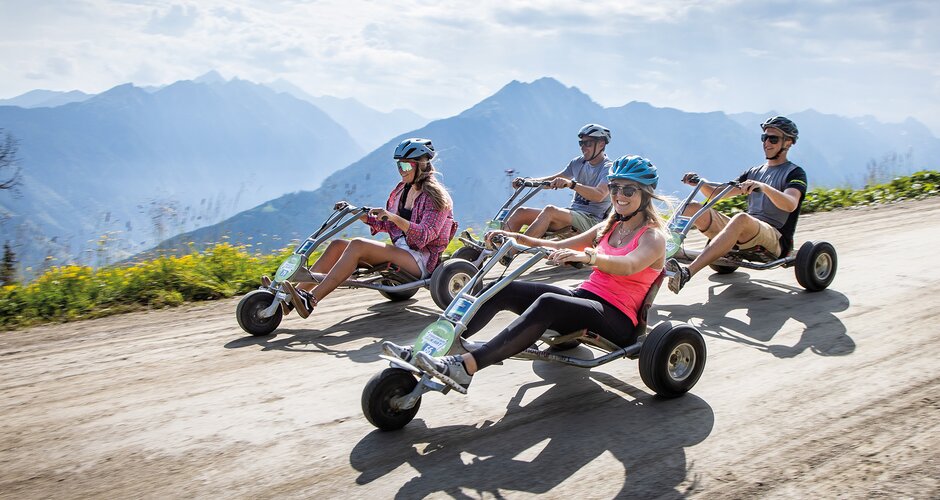 Four helmeted riders ride mountain go-karts along a wide gravel road with mountains in the background | © Planai, Harald Steiner