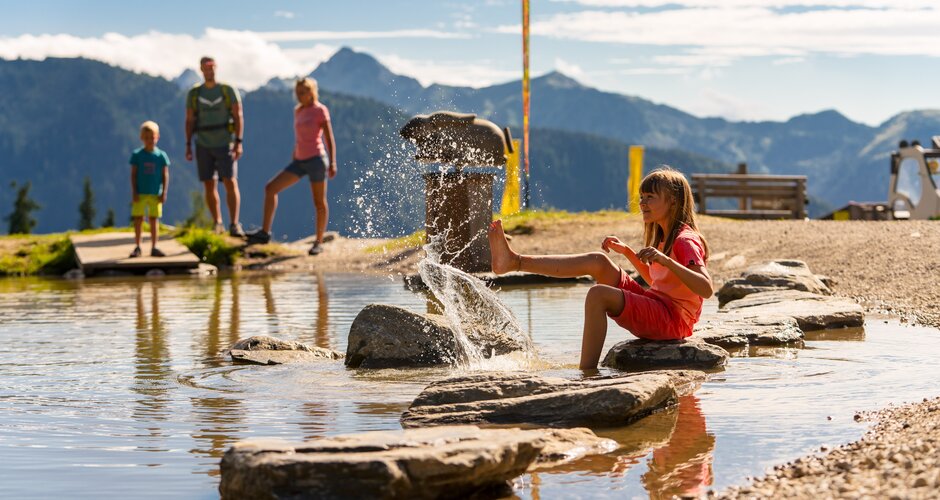 A girl sits on stepping stones in shallow water and splashes while a family stands before the mountains | © Planai, Christine Höflehner