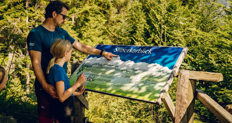 A man points at a wooden panorama board with a girl at the forest edge in Ramsau am Dachstein | © TVB Schladming-Dachstein, Christine Höflehner