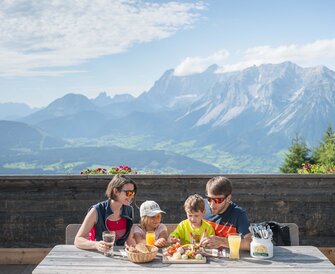 Family with two kids enjoys snack board and drinks at a wooden table on a summer hut terrace with scenic view of the Alps.