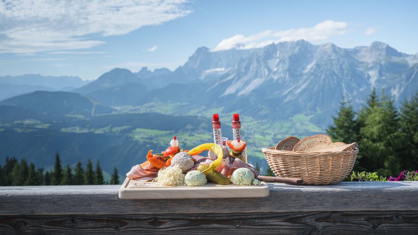 Traditional snack board with cheese, sausage, spreads and bread on a bench with a stunning Alpine panorama behind.
