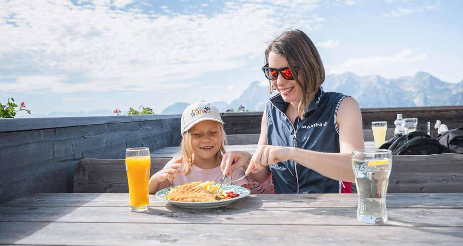 Mother helps child eat schnitzel and fries at a sunny mountain hut table with scenic alpine view in the background.