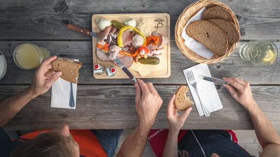 Two people enjoy alpine cold cuts and bread at a rustic wooden table with cheese, meat and drinks from top view.