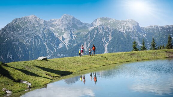 A family stands by a calm mountain lake in sunshine with clear reflection and dramatic alpine backdrop in Ski amadé region.