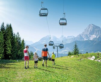 A family hikes across an alpine meadow in the sun, with a view of gondolas in the air and the impressive Dachstein mountains beyond.