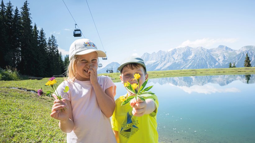 Two kids holding picked flowers at a mountain lake in Ski amadé, with the striking Dachstein mountains in the background.