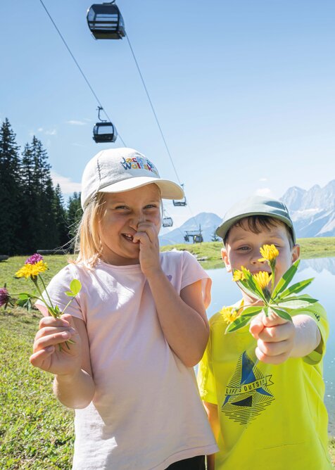 Two kids holding picked flowers at a mountain lake in Ski amadé, with the striking Dachstein mountains in the background.