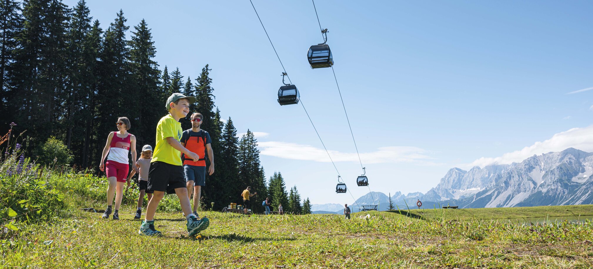 A family hikes across an alpine meadow beneath gondolas, enjoying the sunshine and mountain views in the Ski amadé region.