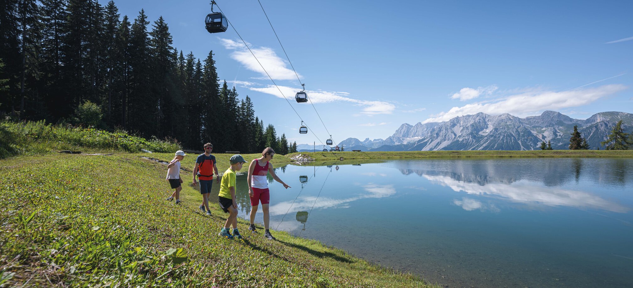 A family walks along a crystal-clear mountain lake under gondolas, enjoying the stunning view of the Dachstein mountains.