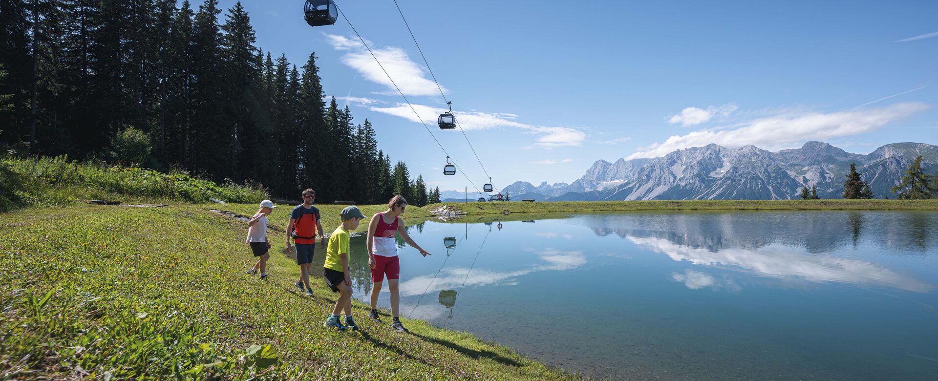 Eine Familie spaziert entlang eines spiegelklaren Bergsees unter schwebenden Gondeln mit Aussicht auf das Dachsteinmassiv.