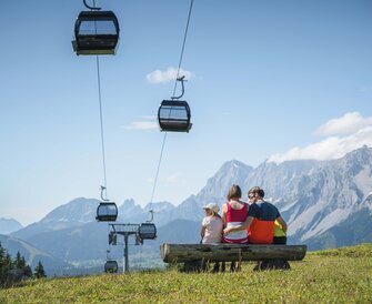 A family sits on a wooden bench under gondolas, admiring the impressive mountain panorama in the Austrian region of Ski amadé.