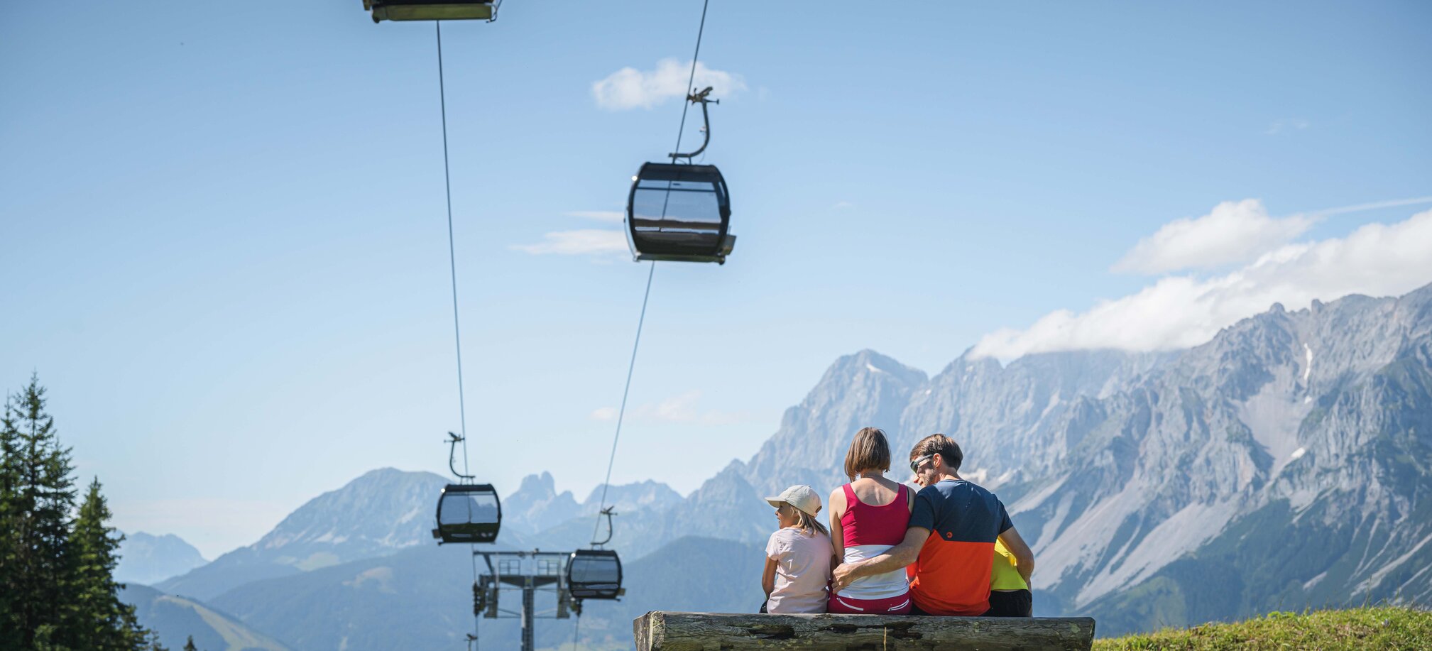 A family sits on a wooden bench under gondolas, admiring the impressive mountain panorama in the Austrian region of Ski amadé.