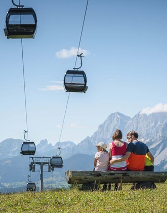A family sits on a wooden bench under gondolas, admiring the impressive mountain panorama in the Austrian region of Ski amadé.