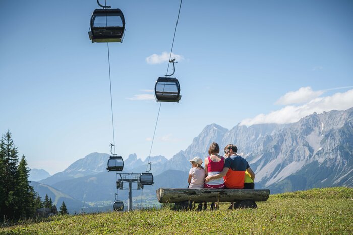 A family sits on a wooden bench under gondolas, admiring the impressive mountain panorama in the Austrian region of Ski amadé.