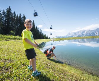 A smiling boy plays at the edge of a clear mountain lake while his father kneels behind him, gondolas float above the scenery.