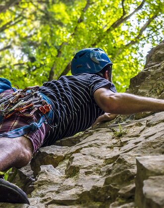 A climber with helmet and full gear ascends a steep rock face surrounded by forest, bathed in warm summer light.