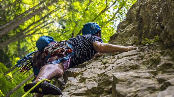 A climber with helmet and full gear ascends a steep rock face surrounded by forest, bathed in warm summer light.