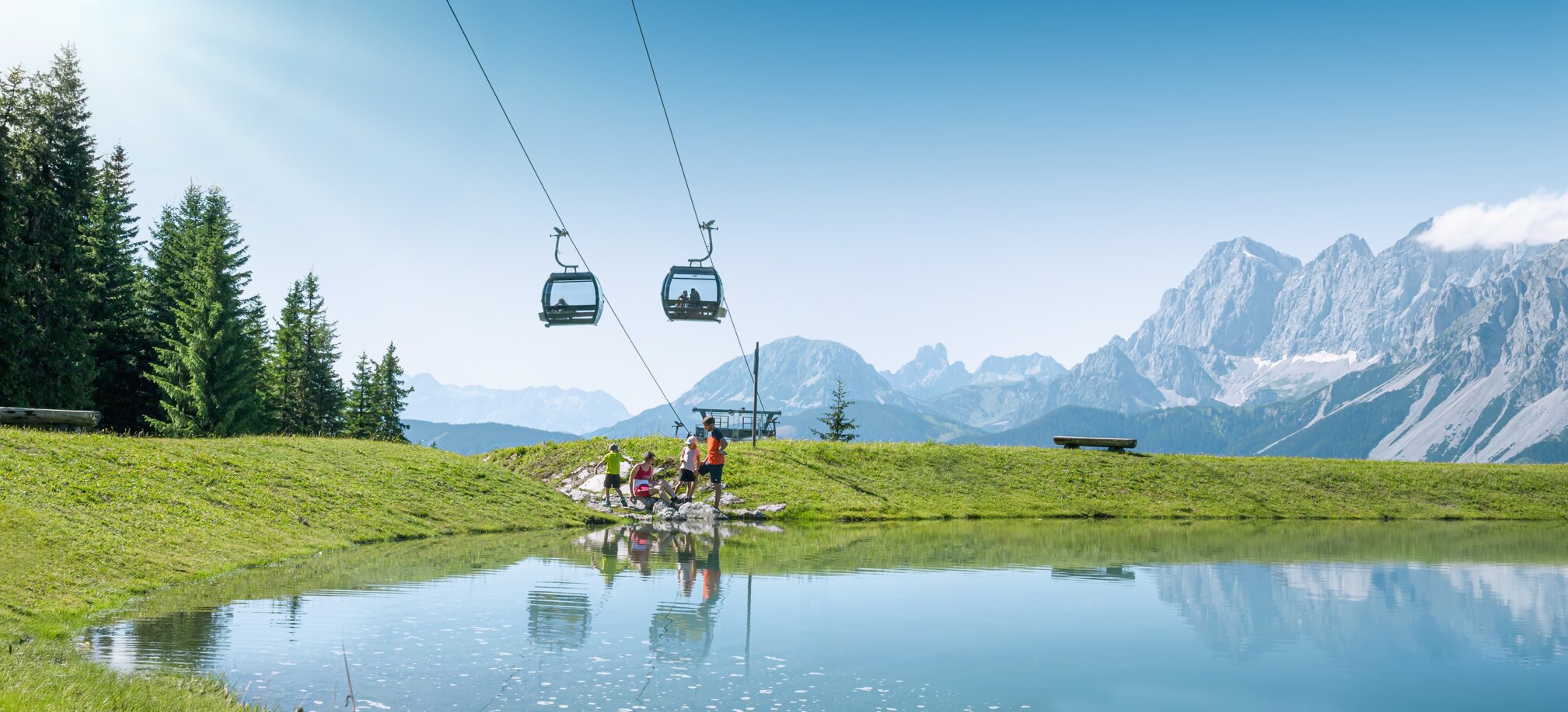 Family by the shore of a small mountain pond beneath a lift, with green meadows and Alps behind