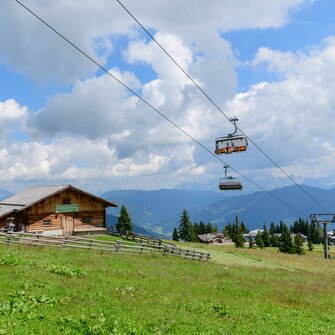 Two rustic alpine huts on a green meadow with a passing chairlift under a blue sky with scattered clouds.