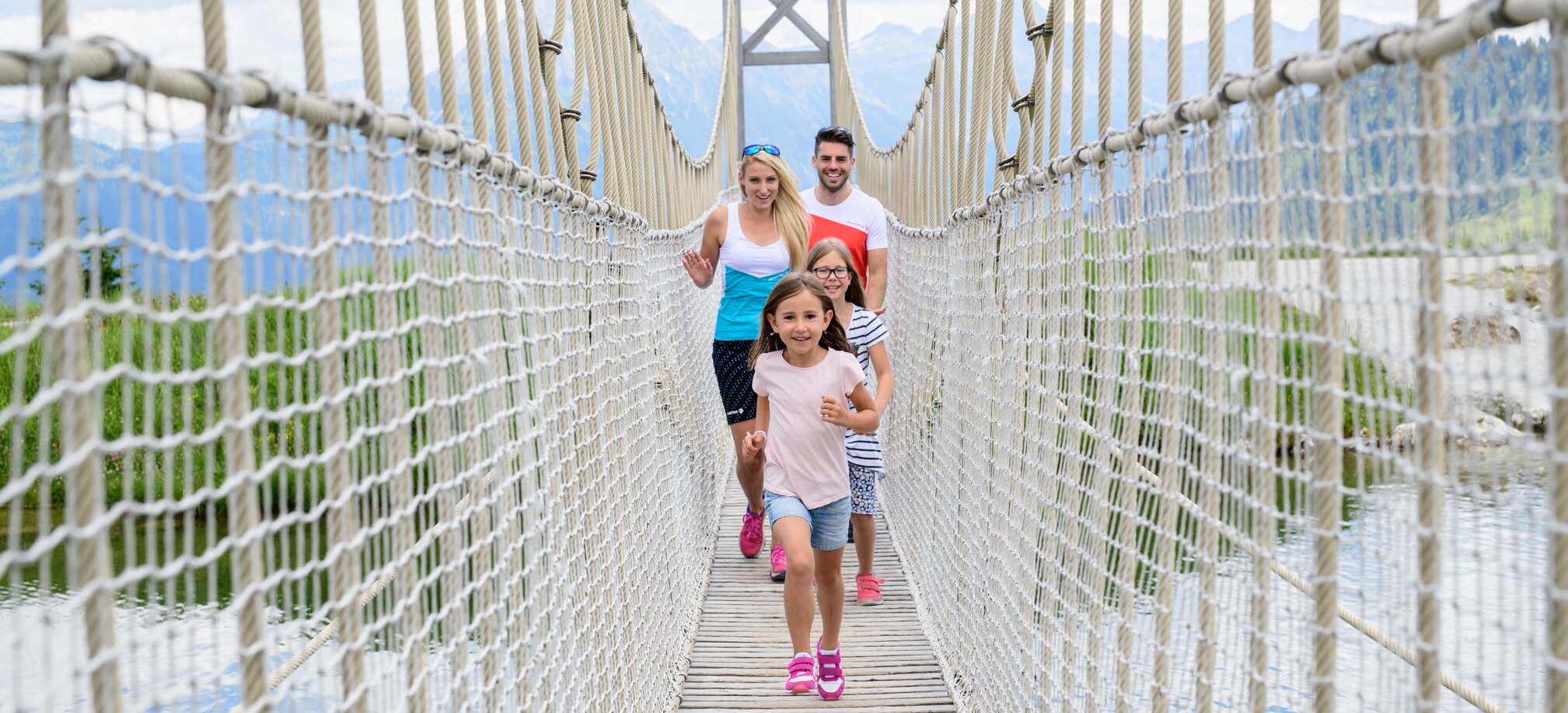 Two children and their parents crossing a rope suspension bridge during a summer family hike in the alpine mountains.