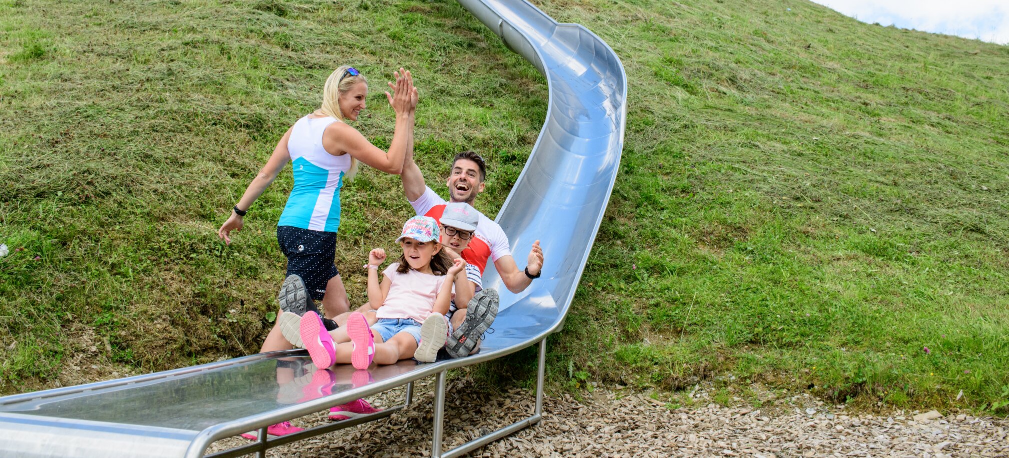 Two kids and their parents laugh while sliding down a long metal slide in the mountains – a joyful alpine family moment.