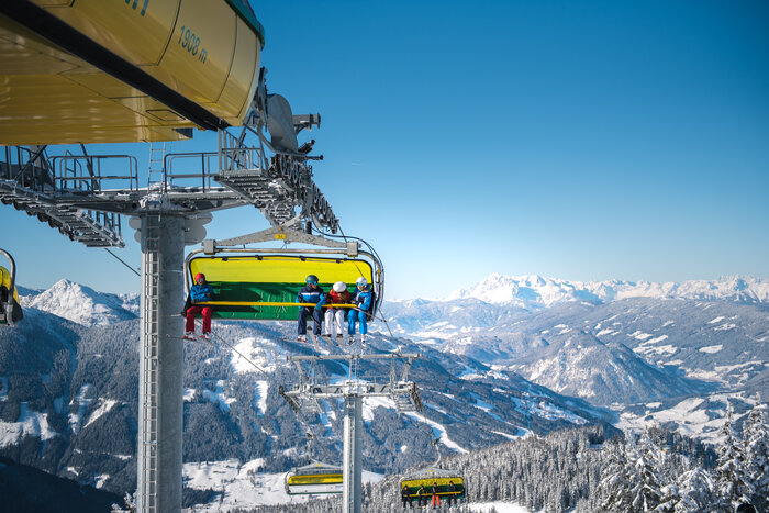 Skiers in colorful ski suits ride the chairlift, with a snowy Alpine mountain panorama in the background.