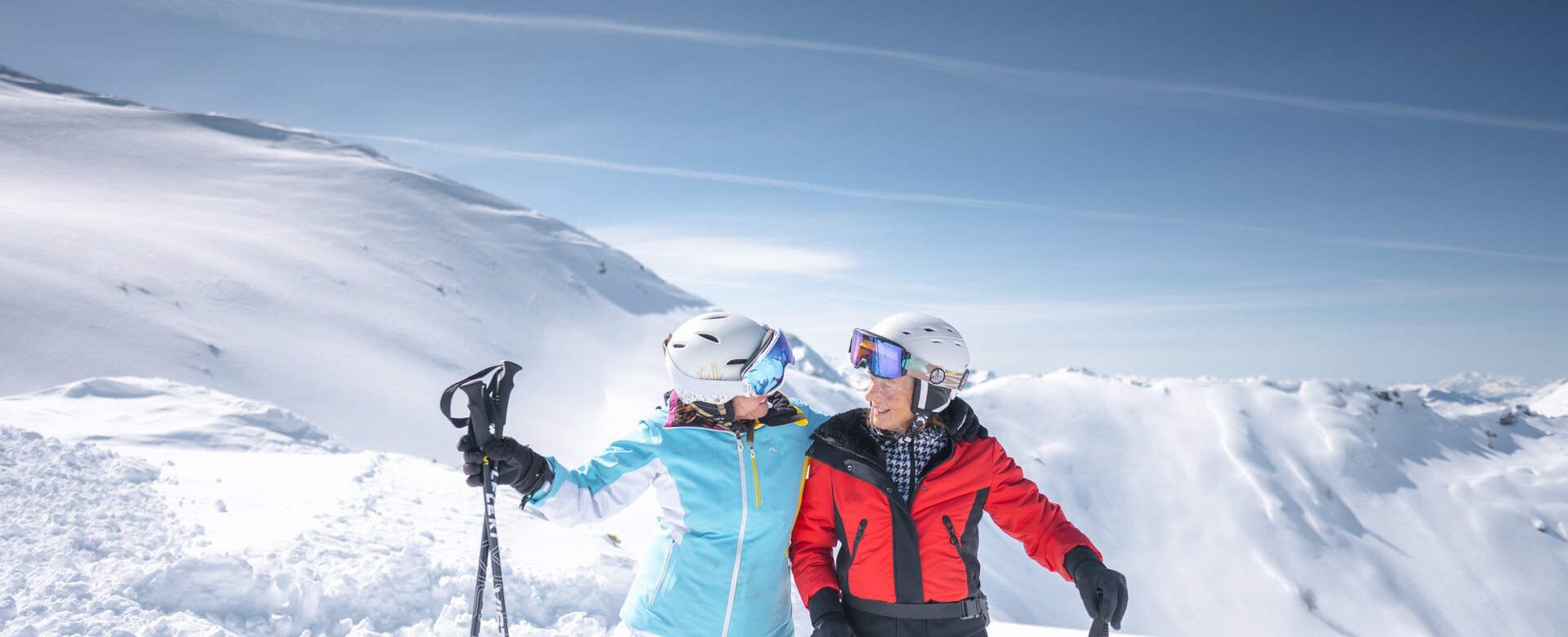Two best ager female skiers hug and laugh on the slope, surrounded by snowy mountains, holding ski poles.