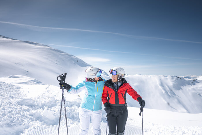 Two best ager female skiers hug and laugh on the slope, surrounded by snowy mountains, holding ski poles.