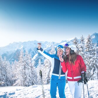 Two female skiers take a selfie at the piste edge with snowy Alpine backdrop, smiling and enjoying the sun.