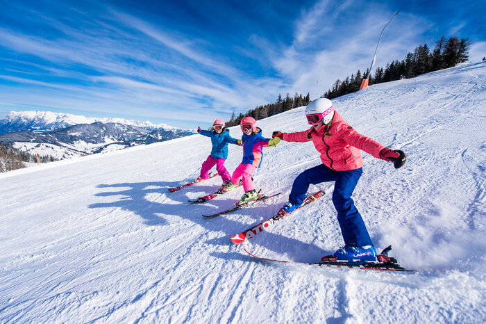 Three kids in colorful ski outfits ski side by side on the slope, holding hands and laughing in the snow.