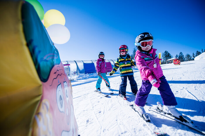 Three kids in colorful ski suits ski on a sunny slope between ski school features, smiling and motivated.