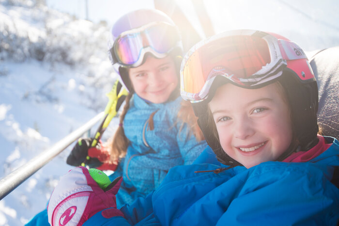 Two kids in colorful ski suits and helmets sit smiling on the ski lift, enjoying the sun and snowy landscape.