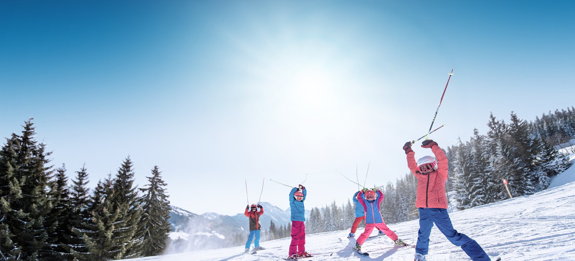 Kids in colorful ski outfits raise ski poles happily in the air, with snowy mountains and slopes in the background.