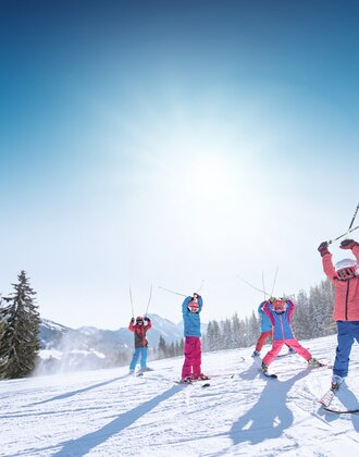 Kids in colorful ski outfits raise ski poles happily in the air, with snowy mountains and slopes in the background.