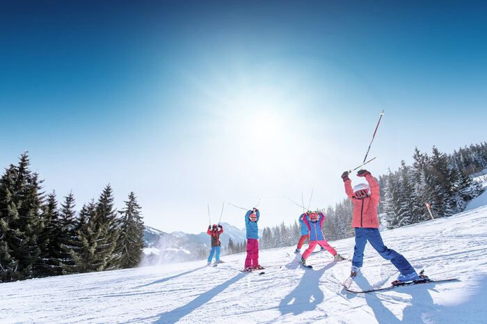 Kids in colorful ski outfits raise ski poles happily in the air, with snowy mountains and slopes in the background.