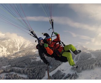 Two people soar through the sky during tandem paragliding over a snow-covered mountain landscape, surrounded by a winter panorama.