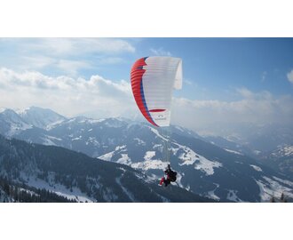 Paraglider with red, blue and white glider flies over mountains covered in forest and snowy pistes