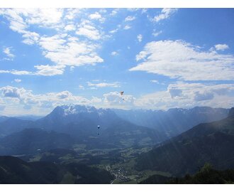Several paragliders soar high above a green alpine landscape with mountains and valleys under a blue sky. | © Flugschule Aufwind