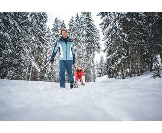 A man pulls a laughing woman on a sled along a snowy forest path. | © Ski amadé