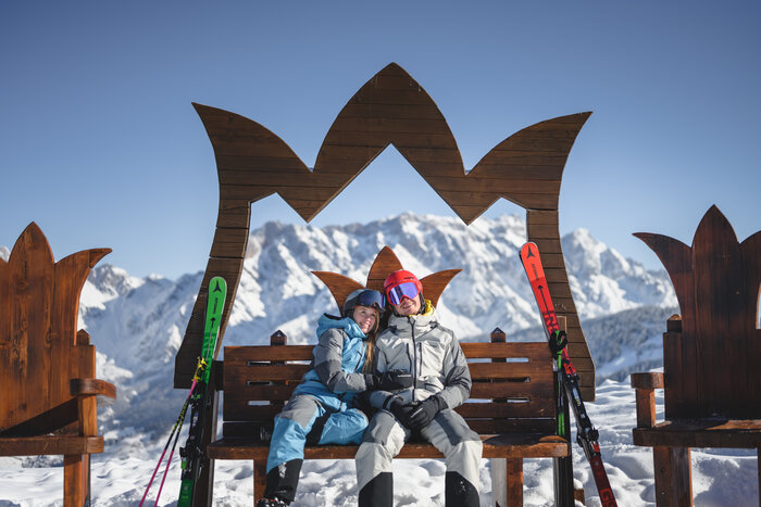 Two people in ski outfits sit on a wooden bench with a crown frame & marvel at the panorama | © Christian Schartner