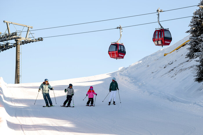 Four skiers, including two children in colorful outfits, skiing on a groomed slope, with red gondolas in the background. | © Gerhard Wolkersdorfer