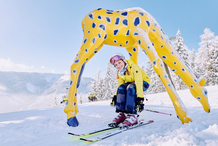 Skier in yellow jacket skiing through a yellow giraffe figure, snow-covered, blue sky, mountains in the background. | © Bergbahnen Filzmoos