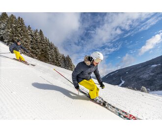Two skiers, each wearing yellow trousers and a grey jacket, take the bend to ski down the piste