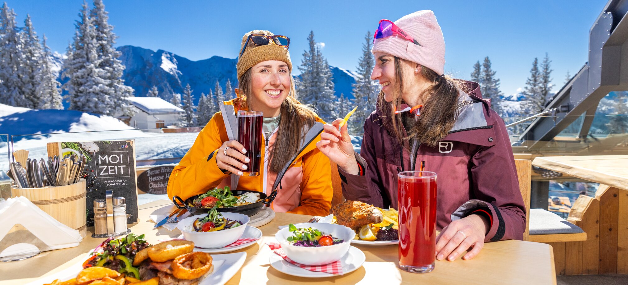 Two women eat and drink in the sun on the terrace with snowy mountain views. | © Hauser Kaibling