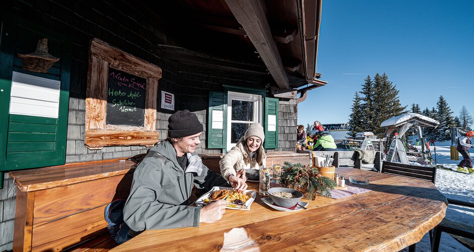 Two people eat outside at a ski hut with wooden bench and snowy background. | © Hauser Kaibling